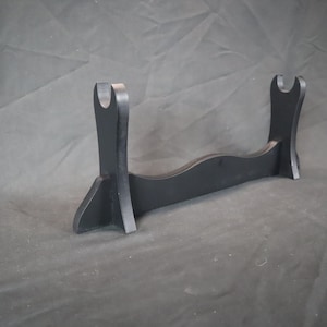 Peut inclure: Un pr&eacute;sentoir &agrave; &eacute;p&eacute;e en bois noir, avec une base incurv&eacute;e et deux supports verticaux avec des d&eacute;coupes en forme de U. Con&ccedil;u pour contenir une &eacute;p&eacute;e ou un article similaire. Le support mesure environ 30 cm de large.