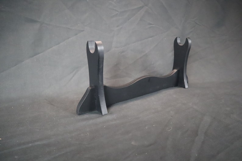 Peut inclure: Un pr&eacute;sentoir &agrave; &eacute;p&eacute;e en bois noir, avec une base incurv&eacute;e et deux supports verticaux avec des d&eacute;coupes en forme de U. Con&ccedil;u pour contenir une &eacute;p&eacute;e ou un article similaire. Le support mesure environ 30 cm de large.