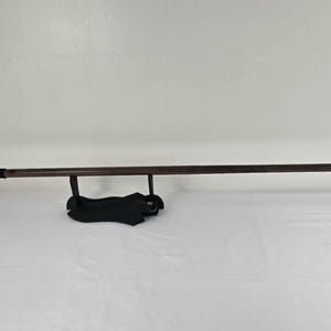 May include: A long spear with a dark brown wooden shaft and a black and silver spearhead. The spear is displayed on a black stand against a white background. The spearhead has a decorative design.