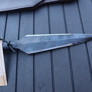 May include: A hand-forged spear with a black leather sheath. The spear has a long, pointed blade and a wooden handle. The tip of the spear has been quenched and hardened. Expect discoloration, chat, and or a tort C.