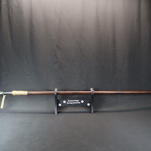 May include: A brown wooden spear with a metal tip and a brown leather wrap. The spear is mounted on a black stand with the text "Guardian Dragon The Holy Sword Shop" printed on it.