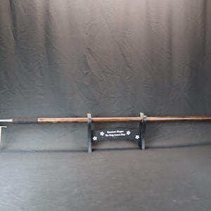 May include: A long spear with a silver blade and a dark wooden shaft. The handle is wrapped in black material. The spear is displayed on a black stand with white floral accents. The text on the stand reads "Quantum Shoppe The Holy Sword Shop".