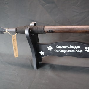 May include: A black wooden stand with a dark brown spear resting on it. The stand has white cherry blossoms and the text "Quantum Shoppu The Only Isekai Shop" on it.