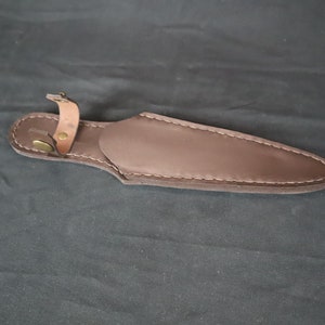 May include: A brown leather knife sheath with a stitched edge and a strap with a brass button closure. The sheath is designed to hold a knife and protect the blade.