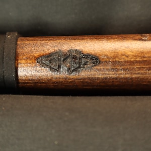 Peut inclure: Gros plan sur un objet en bois avec une finition marron fonc&eacute;. Une poign&eacute;e en caoutchouc noir est visible pr&egrave;s du haut. Le bois pr&eacute;sente un motif sombre grav&eacute; pr&egrave;s du centre. L'objet semble &ecirc;tre une poign&eacute;e ou une partie d'un outil plus grand.