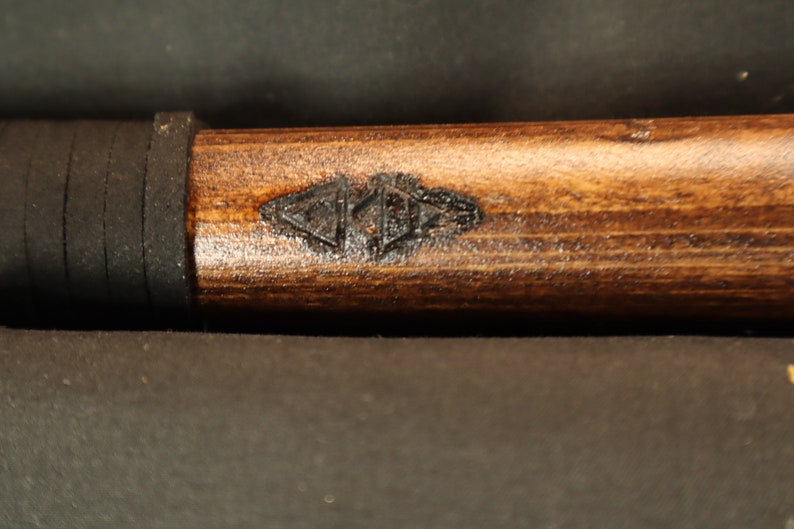 Peut inclure: Gros plan sur un objet en bois avec une finition marron fonc&eacute;. Une poign&eacute;e en caoutchouc noir est visible pr&egrave;s du haut. Le bois pr&eacute;sente un motif sombre grav&eacute; pr&egrave;s du centre. L'objet semble &ecirc;tre une poign&eacute;e ou une partie d'un outil plus grand.