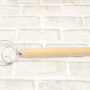 May include: A wooden handled dough whisk with a metal loop and a double spiral design. The whisk is on a white brick background.
