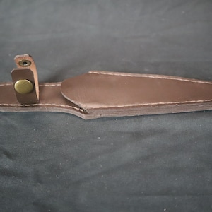 Peut inclure: Un &eacute;tui &agrave; couteau en cuir marron avec des coutures blanches et une fermeture &agrave; bouton en laiton. L'&eacute;tui est con&ccedil;u pour contenir un couteau et poss&egrave;de une boucle pour le fixer. Le cuir est d'un brun fonc&eacute; riche.