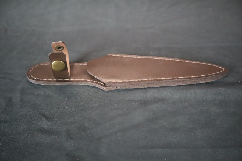 Peut inclure: Un &eacute;tui &agrave; couteau en cuir marron avec des coutures blanches et une fermeture &agrave; bouton en laiton. L'&eacute;tui est con&ccedil;u pour contenir un couteau et poss&egrave;de une boucle pour le fixer. Le cuir est d'un brun fonc&eacute; riche.