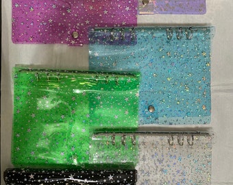May include: A selection of clear wallets with a binder mechanism, in shades like purple and green. Each wallet has a star pattern and a silver metal ring binder. The wallets have a snap closure.