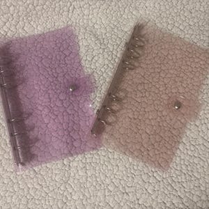 May include: Two clear binders with silver metal rings and snap closures. One is light purple, the other light peach. The binders have a textured finish. They are on a white, textured surface.