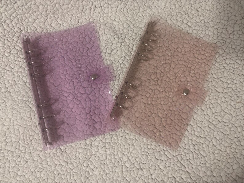 May include: Two clear binders with silver metal rings and snap closures. One is light purple, the other light peach. The binders have a textured finish. They are on a white, textured surface.