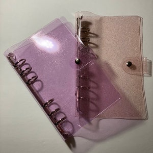 May include: Two clear, glittery binders. One is a light purple colour, and the other is a light pink colour. Both binders have silver metal rings and a snap closure. The binders are on a white surface.