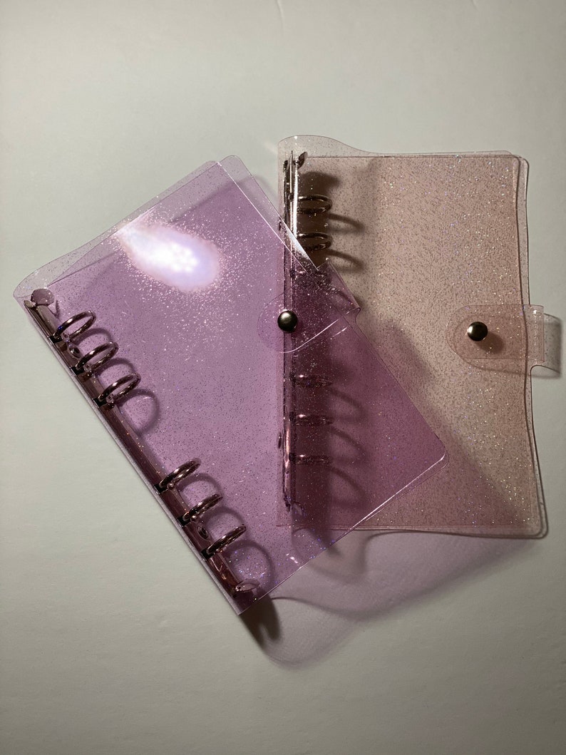 May include: Two clear, glittery binders. One is a light purple colour, and the other is a light pink colour. Both binders have silver metal rings and a snap closure. The binders are on a white surface.