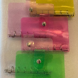 May include: Four clear plastic binders with glitter embedded in the plastic. The binders are stacked on top of each other. The top binder is yellow, the next is orange, the next is pink, and the bottom binder is green.