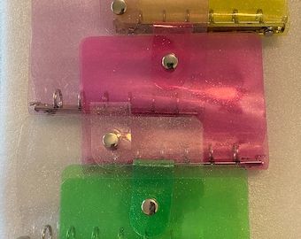 May include: Four clear plastic binders with glitter embedded in the plastic. The binders are stacked on top of each other. The top binder is yellow, the next is orange, the next is pink, and the bottom binder is green.