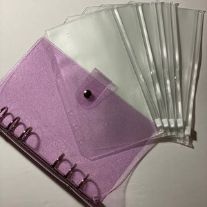 May include: A clear, glittery purple binder with silver rings and a press stud closure. Several clear plastic envelope-style pouches are fanned out beside the binder. The binder and pouches are designed for organizing and storing items.