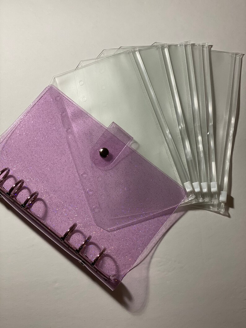 May include: A clear, glittery purple binder with silver rings and a press stud closure. Several clear plastic envelope-style pouches are fanned out beside the binder. The binder and pouches are designed for organizing and storing items.
