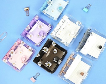May include: A selection of clear mini binders, adorned with daisy patterns, in shades of blue, purple, pink, and black. Each binder holds lined paper and features a snap closure. Gold and pastel paper clips are arranged around the binders on a blue surface.