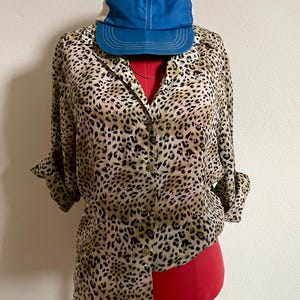 Puede incluir: Una camisa de botones de manga larga con estampado de leopardo marrón y beige con una gorra de béisbol azul en la parte superior.