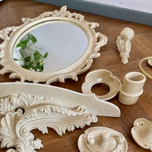 Puede incluir: Un conjunto de accesorios de baño de cerámica de color crema con un diseño floral ornamentado. El conjunto incluye un espejo, un estante, un portacepillos de dientes, un jabonera y dos ganchos.