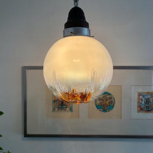 May include: A vintage pendant light fixture with a frosted glass globe. The globe has a clear, textured bottom with amber-colored accents. The fixture hangs from a silver chain and has a black and silver hardware.