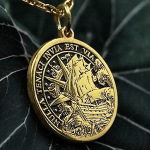 May include: Gold necklace with a round pendant featuring a ship sailing through rough waves. The pendant has the inscription "Nulla Invia Est Via" around the edge.