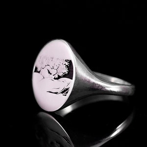 May include: A silver signet ring with a round, engraved top featuring a detailed design. The ring has a smooth, polished finish.