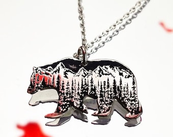 Pode incluir: Um colar de prata com um pingente de urso. O urso é contornado em preto e tem uma cena de montanha dentro. O colar está sobre um fundo branco.