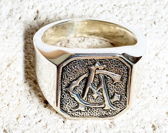 May include: Two signet rings, one gold and one silver, with a monogram design. The rings are on a white textured surface.