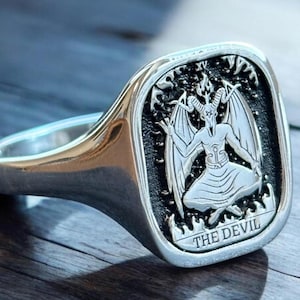 May include: A silver signet ring with a square face featuring a black and white image of the Devil card from a tarot deck. The card reads "THE DEVIL".