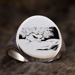May include: A silver signet ring with an engraved image of a couple embracing. The ring is oval shaped and has a smooth, polished finish.