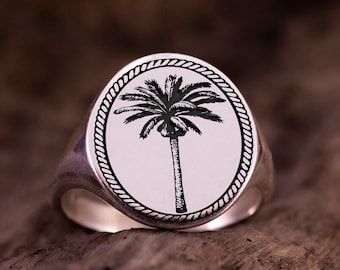 May include: Silver signet ring with an oval face. It features a black palm tree design within a rope-like border. The ring has a polished finish and a classic style. The ring is shown on a wooden surface.