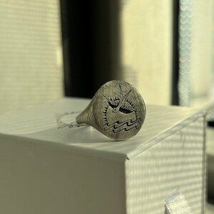 May include: A silver signet ring with an engraved design. The ring is in a white box with a white ribbon.