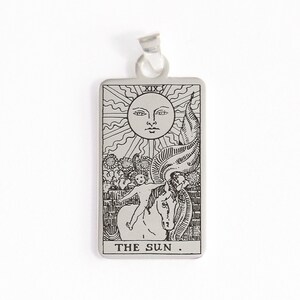 May include: A silver pendant featuring a tarot card design. The card depicts the Sun card, with the Roman numeral XIX at the top and the words "THE SUN." at the bottom.