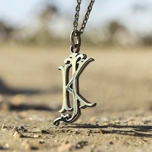 以下が含まれることがあります： 装飾的な文字Kのチャームが付いたシルバーのペンダントネックレス。ペンダントはシルバーチェーンにぶら下がっており、砂地の表面に置かれています。