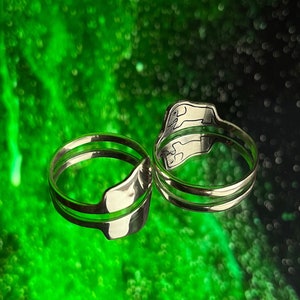 Puede incluir: Dos anillos de plata con un dise&ntilde;o &uacute;nico, uno de los anillos es ligeramente m&aacute;s grande que el otro, ambos anillos est&aacute;n sobre un fondo verde y negro.