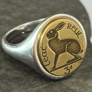 Op de afbeelding: Zilveren ring met een goudkleurige, ronde voorkant. De voorkant toont een gedetailleerde illustratie van een haas en de tekst "Lear" en "Reul". De ring heeft een klassieke zegelringstijl.