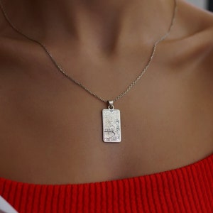 Op de afbeelding: Een zilveren ketting met een rechthoekige hanger met een tarotkaartontwerp. De kaart toont De Geliefden, een grote arcana-kaart in het tarotdeck.