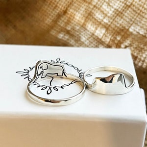 Puede incluir: Tres anillos de plata en una caja blanca. Los anillos son de dise&ntilde;o simple y moderno. Un anillo presenta una silueta de elefante.