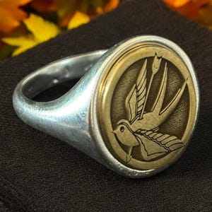 May include: A silver ring with a circular, gold-colored face featuring a detailed engraving of a bird in flight. The ring is set against a dark background, with blurred autumn foliage in the background.