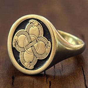 Peut inclure: Une bague chevalière dorée avec une face ovale. La face présente un motif symétrique détaillé en or sur un fond noir. Le corps de la bague est lisse et poli, reflétant la lumière. Le motif semble être un nœud celtique.