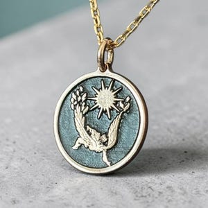 May include: Gold-toned necklace featuring a circular pendant with a teal background. The pendant showcases a sunburst design with a bird-like figure and wheat stalks. The necklace is a delicate piece of jewelry.