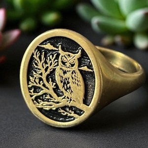 May include: A gold-colored ring with an oval face featuring a detailed owl design. The owl is perched on a branch against a black background. The ring has a smooth, polished finish and a classic signet style.