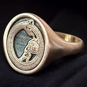 May include: A gold-toned ring featuring an ouroboros design, a serpent eating its own tail, within an oval frame. The ring has a polished finish and a detailed serpent motif. The background is a dark, solid color.