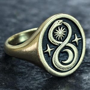 May include: A gold-toned signet ring with an oval face. The face features a black background with a snake eating its tail, a sun, and two stars. The ring has a polished finish.
