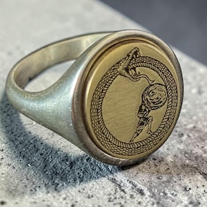 May include: A silver ring with a gold-coloured circular face. The face features a detailed engraving of a snake eating its tail, encircling a figure. The ring has a smooth, polished finish.