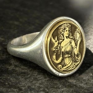 Può includere: Anello color argento con un medaglione ovale dorato raffigurante una figura femminile classica. L'anello presenta un design dettagliato con un'estetica vintage o antica. La figura tiene oggetti, su uno sfondo scuro.