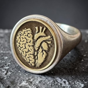 May include: A silver signet ring with a gold-colored circular emblem featuring a detailed brain and heart design. The ring has a polished finish and a rounded band, set against a dark, textured background.