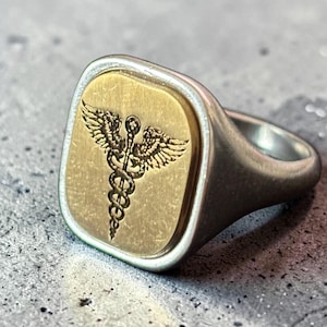 Può includere: Un anello d'argento con una faccia rettangolare color oro. La faccia presenta un'incisione dettagliata del simbolo del Caduceo, un bastone con due serpenti e ali. L'anello ha una finitura in metallo spazzolato.